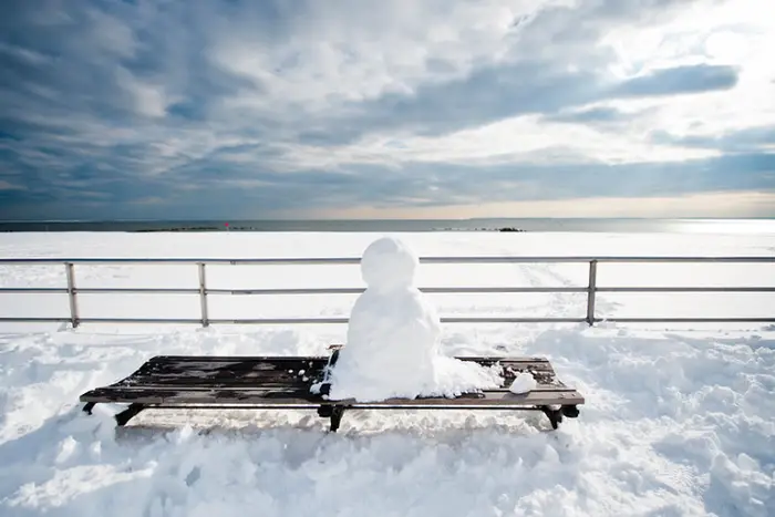 This is a photo of a snowman on a bench at a snow-covered beach.